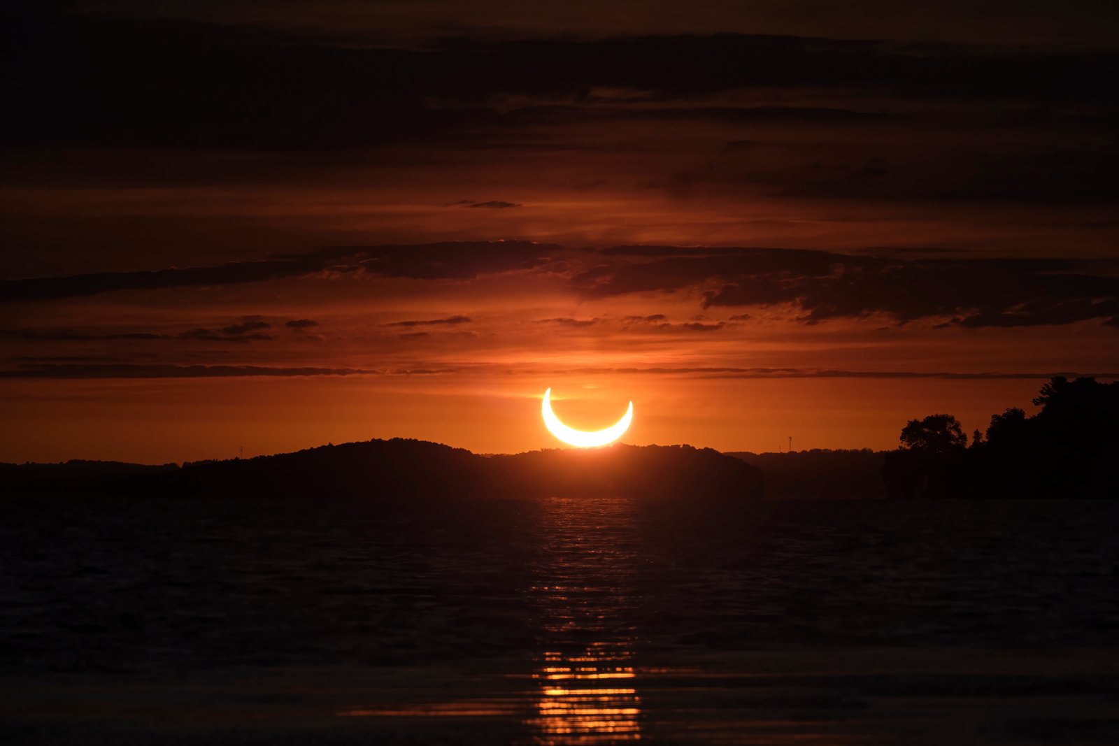 Between layers of clouds, the partial solar eclipse at sunrise is seen over Lake Rice, Ontario, as the sun, partially covered with the moon, in a beautiful orange sky over trees and water.