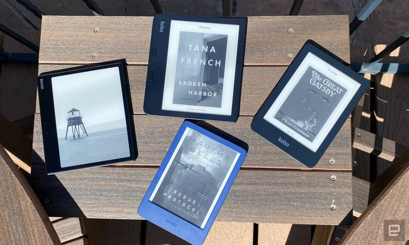 Four ereaders are arranged on a dark brown wooden table outside. There are Boox, Kindle and Kobo devices showing the covers of different novels from Tana French, F. Scott Fitzgerald and Martha Wells.  