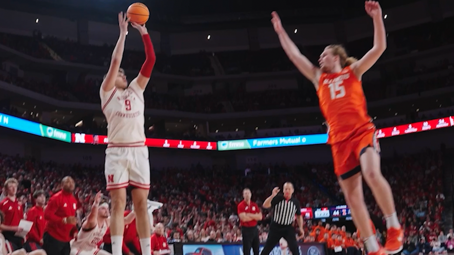 The basketball player shoots the ball while another player tries to ban it.
