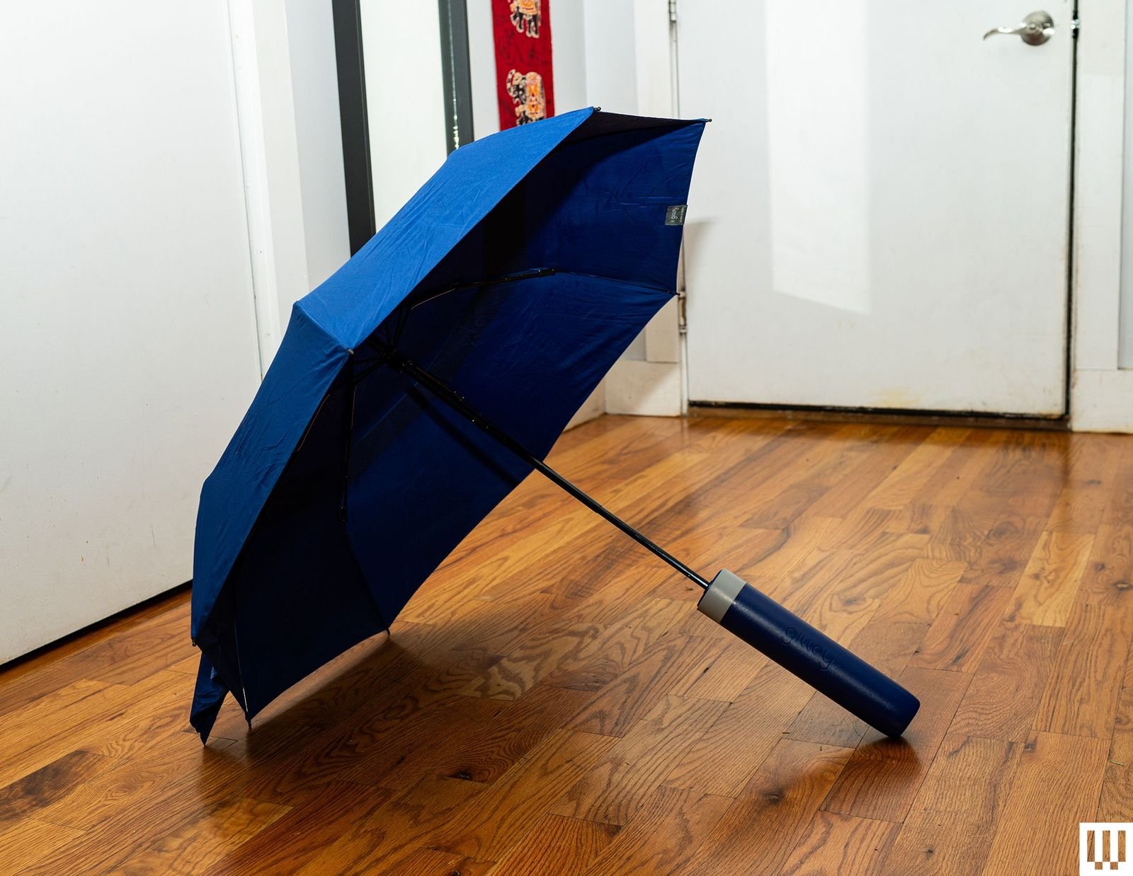 Blue umbrella with sharp edges and a thick handle open and resting on a hardwood floor in a hallway with white walls