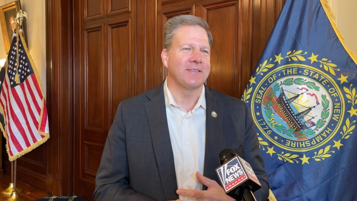 Sununu beside the New Hampshire flag, we know behind it