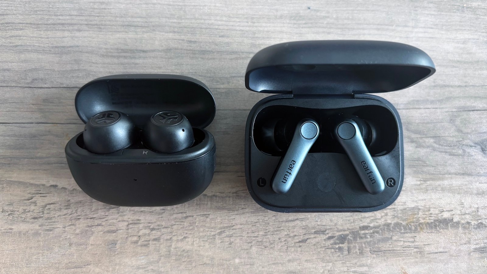 Two black couples of wireless ear headphones, Jlab Go POP AnC and Earfun Air Pro 4, rest on a light brown wooden table with opening their cases, and exposing earphones while shipping inside.