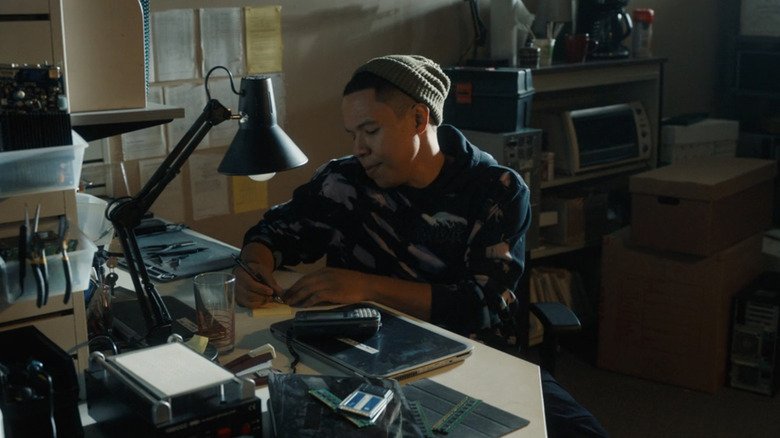 Eric Grace in the role of Bobby Xley sitting on his office writes on a painting in a tracker