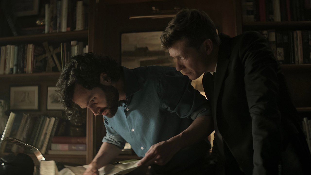 Joe looks at something on a table while Rais hovers behind you.