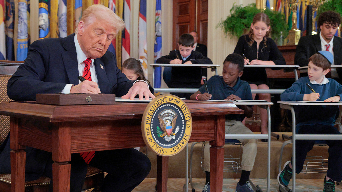 President Donald Trump signs an executive order in an office with children sitting in the offices