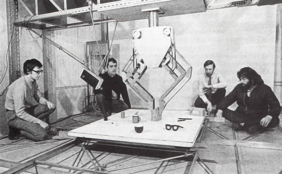 Black and white photo of a robot hovering over a square platform and surrounded by four young men who are crouching as they look at it. 