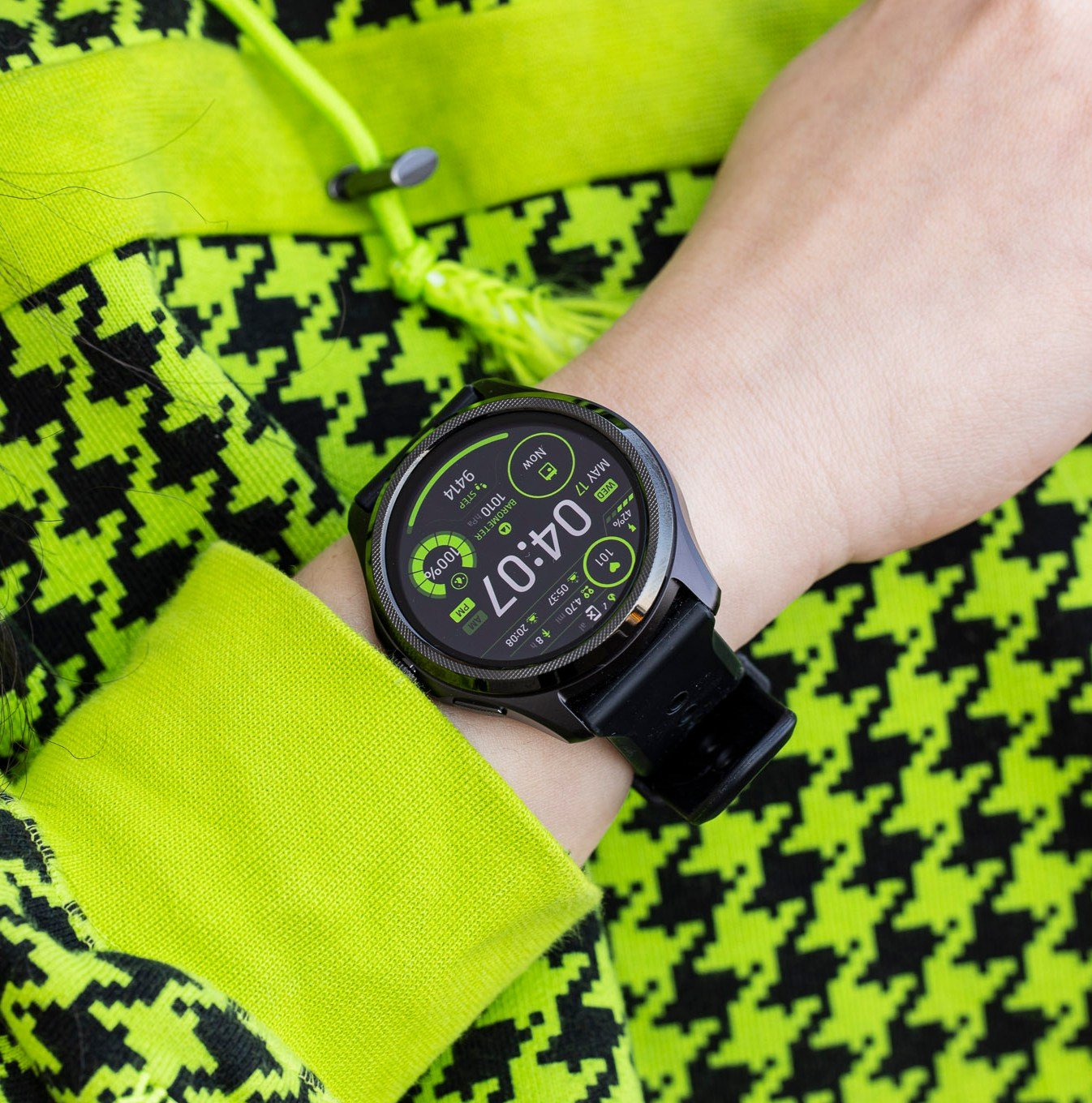 Close-up of the Mobvoi TicWatch Pro 5 on a wrist against a lime green / black sweater with a houndstooth pattern.