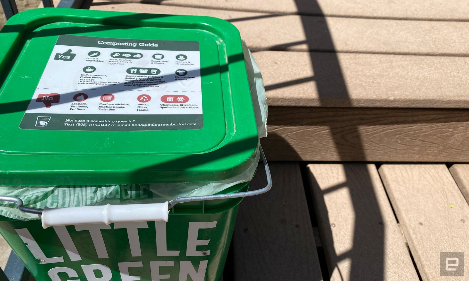 Green and square fertilizer bucket sits in wooden steps. The instructions are listed on foods that can go in the bucket on the cap.