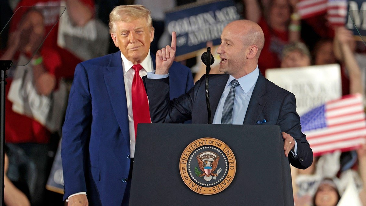 Stephen Miller speaks on a platform with President Donald Trump standing behind and smiling