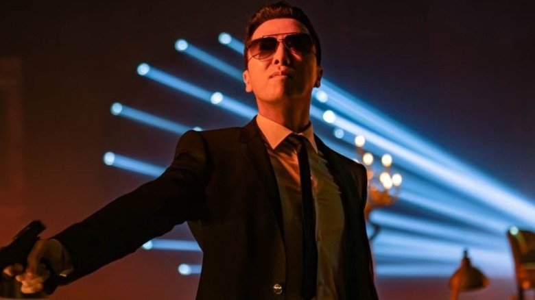Kane from John Wake 4 wears glasses and a suit carrying a gun in front of lead holes in the wall
