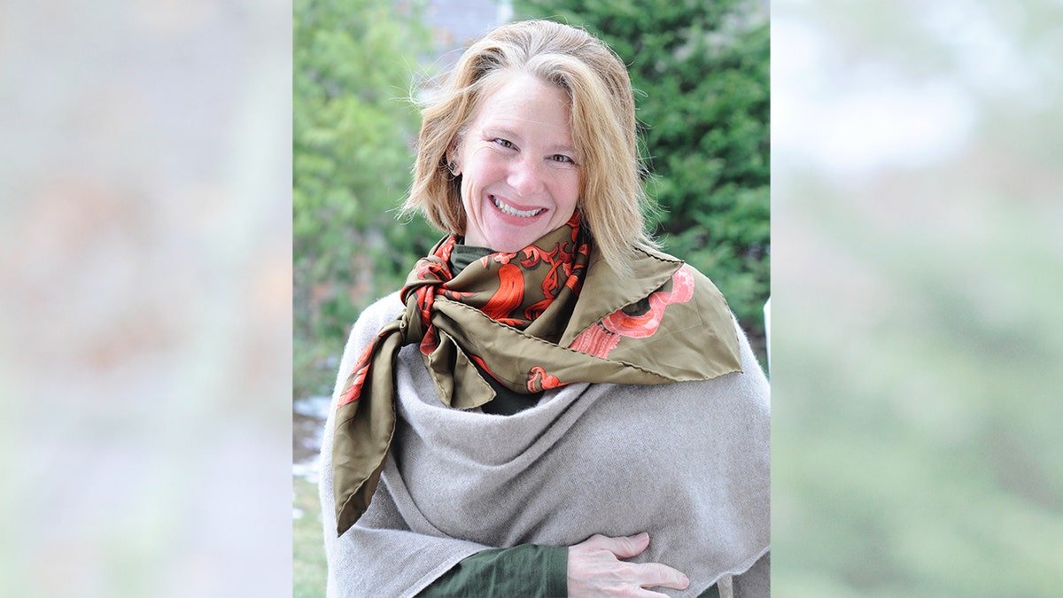 Lisa Brown Langley smiles while wearing a scarf and stands in a wooded area.