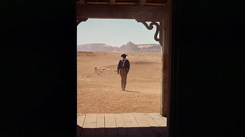Edward Edwards (John Wayne), wears a cowboy hat, walks at the distance towards the deserts, framed with a dark entrance in researchers