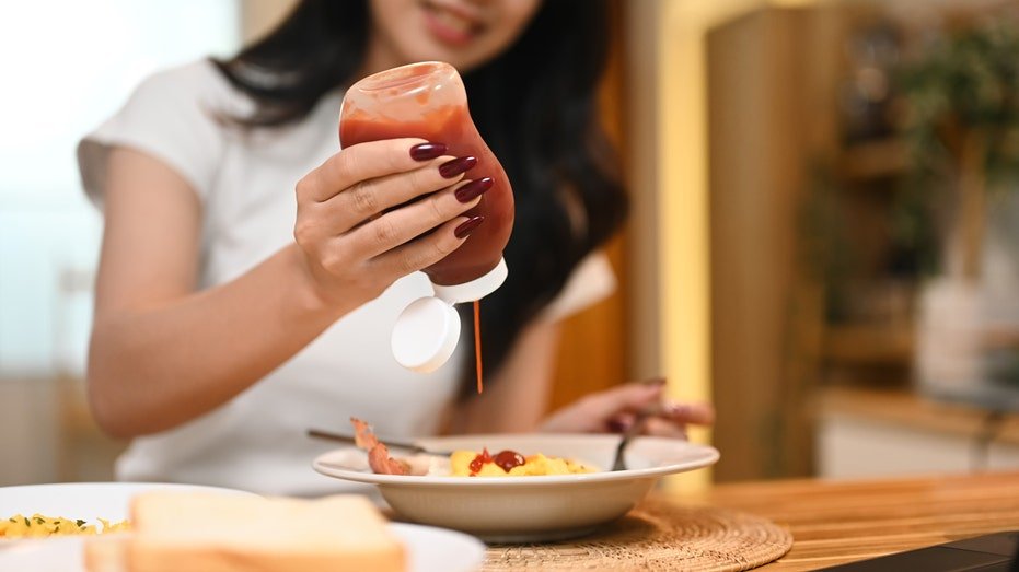 A woman presses ketchup from a bottle on the breakfast dish.