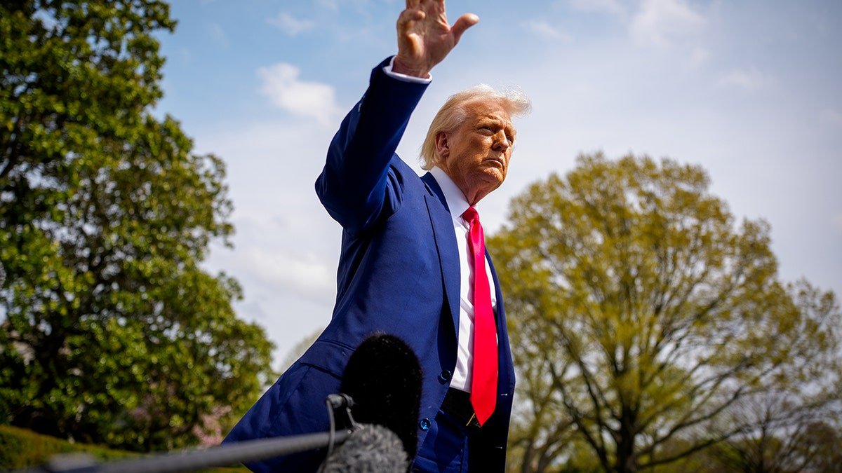 President Donald Trump raises his hand while walking in the open air near the White House, wearing a blue suit and a red tie, with visible trees and microphone at the forefront.