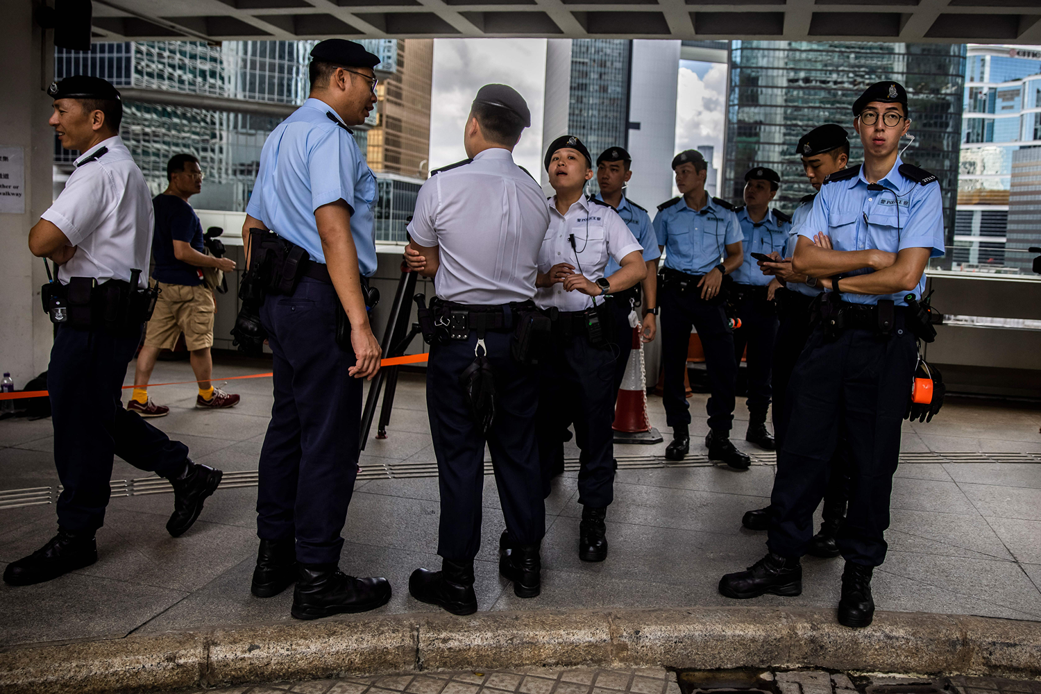 A group of police officers, wearing uniform shirts short sleeves and matching caps, and spoke to each other while they stopped on the sidewalk outside the Hong Kong Supreme Court. Zibing skyscrapers and cloudy sky visible behind officers.