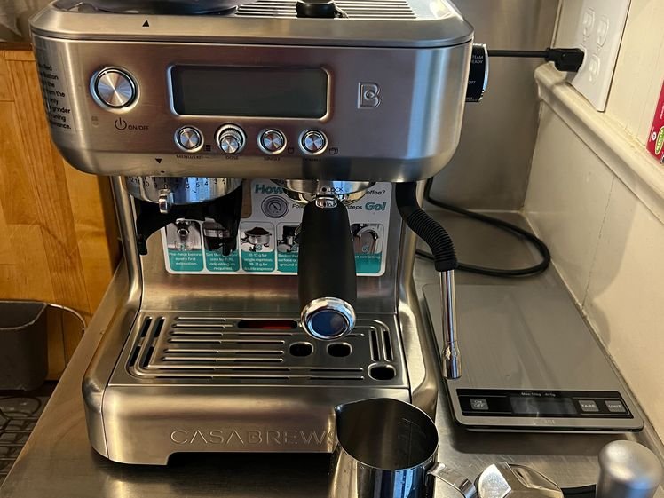 A front view of Casabrews 5700 Pro, which is a silver espresso machine with a small digital screen, sitting on a metal surface with additional supplies next to it