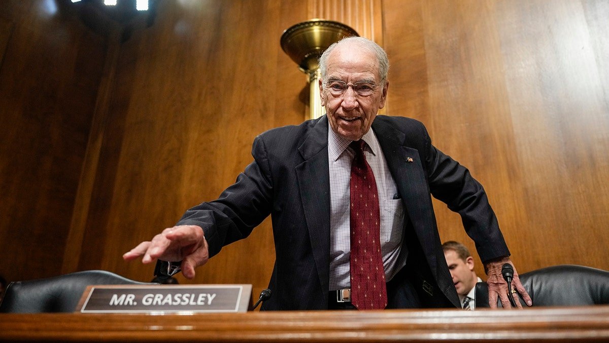 Chuck Grassley in a dark suit, a red tie, reaches the right hand on the table, the left hand on the chair