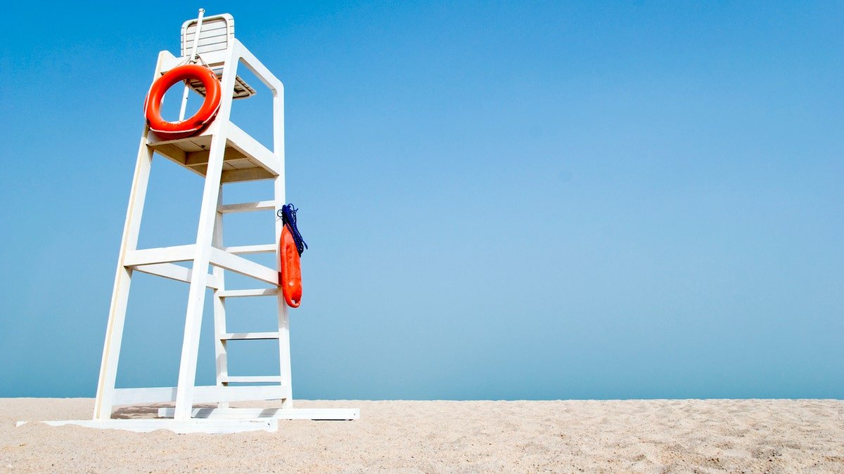 The empty white rescue chair on an empty beach with orange -life buoys hanging on the side. The chair stands against a clear blue sky in the sand.