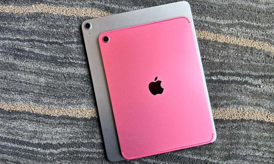 A pink 11-inch Apple iPad rests on top of a silver 13-inch iPad Air. Both tablets are face down on a gray carpet with beige and white streaks.
