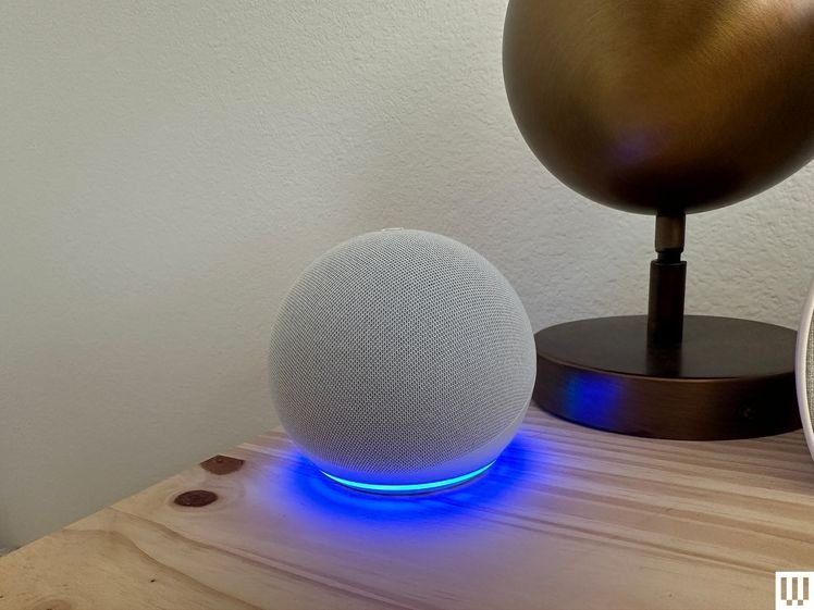 Front view of Echo Dot 5th Generation, a grey sphere device with blue light illuminating from the base while sitting on a wooden nightstand