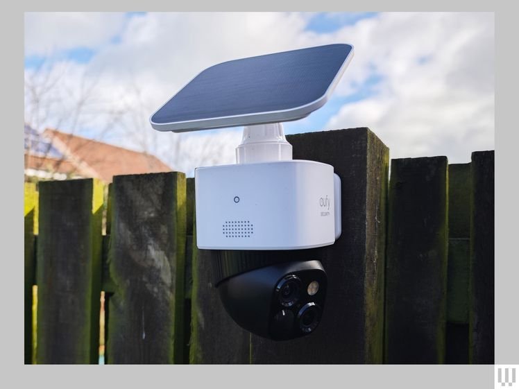 Two part outdoor security camera system attached to a fence. The top is a white box with extended solar panel. The bottom is a black circular set of lenses.