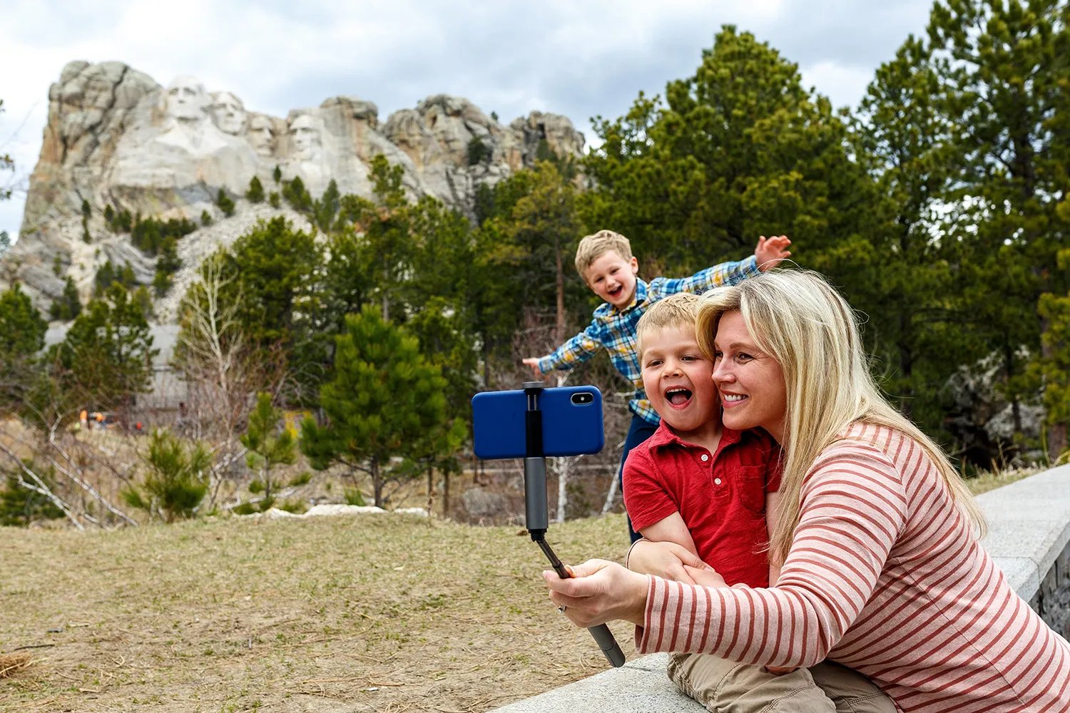 A blond woman uses a striped shirt a personal stick to take a photo with the camera. It carries a small child in a red polo shirt. Another child stands in a shirt engraved behind them, and the arms spread widely while smiling. The Mount Rushmore Memorial monument is waving at the distance, as trees spread between them and the family.