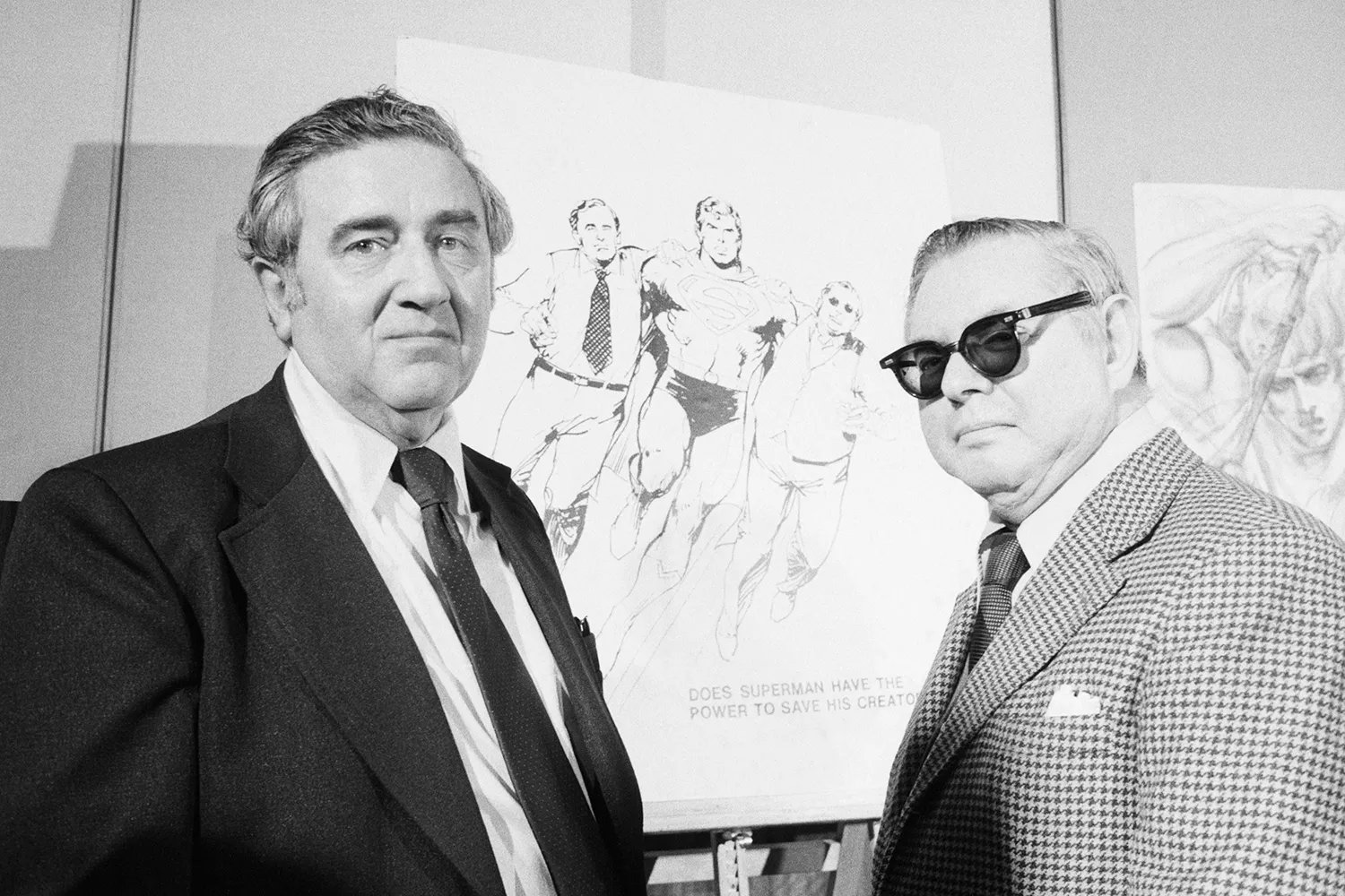 Two men in suits, one wearing sunglasses, post in front of Superman sketches.