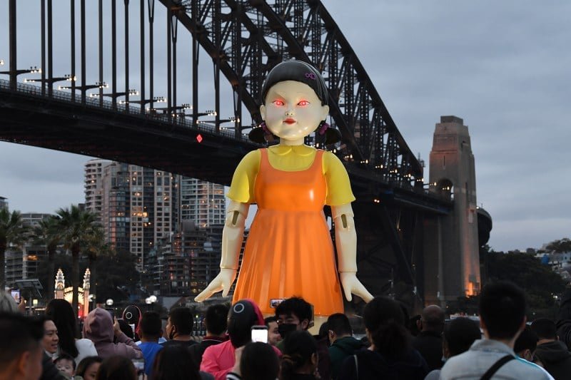 Hundreds of people gather around a giant doll with pigtails, an orange jumper and red glowing eyes.