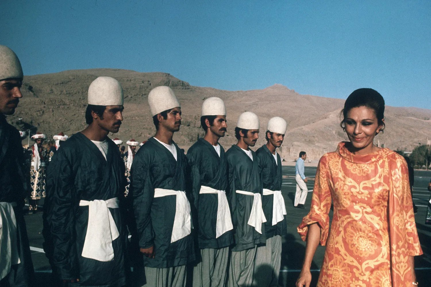 Empress Farah of Iran inspects a row of soldiers dressed in a fashionable 1970s dress.