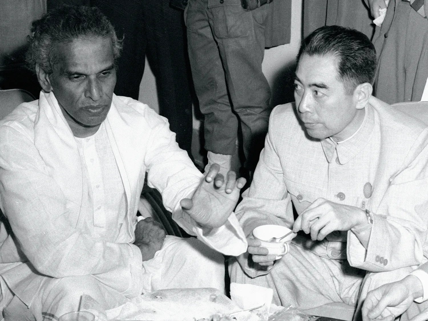In this black-and-white photo, Menon gestures as Zhou drinks from a small cup and leans close to listen. Both men sit cross-legged on the ground with the tea set between them.