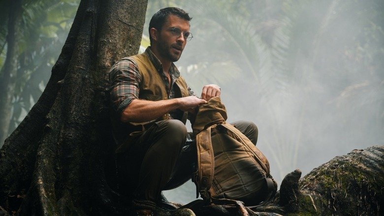 Dr. Henry Lomes sits on a tree with his bag in front of him on the ground in the birth of the global Jurassic