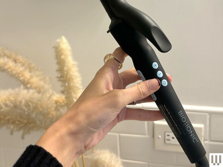 Side view of a hand holding the Bio Ionic Long Barrel Styler, a long narrow black curling iron with three 3 small light blue buttons