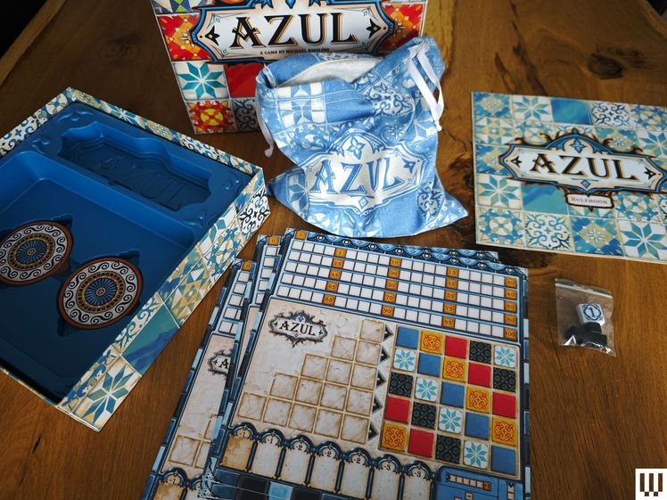 Drawing-themed board game contents scattered on wooden surface. Back: Blue and white box with illustration of a person drawing. Left: Packaging with more supplies and instructions. Right: Spiral notebooks, cards, a sand timer, and a pen.