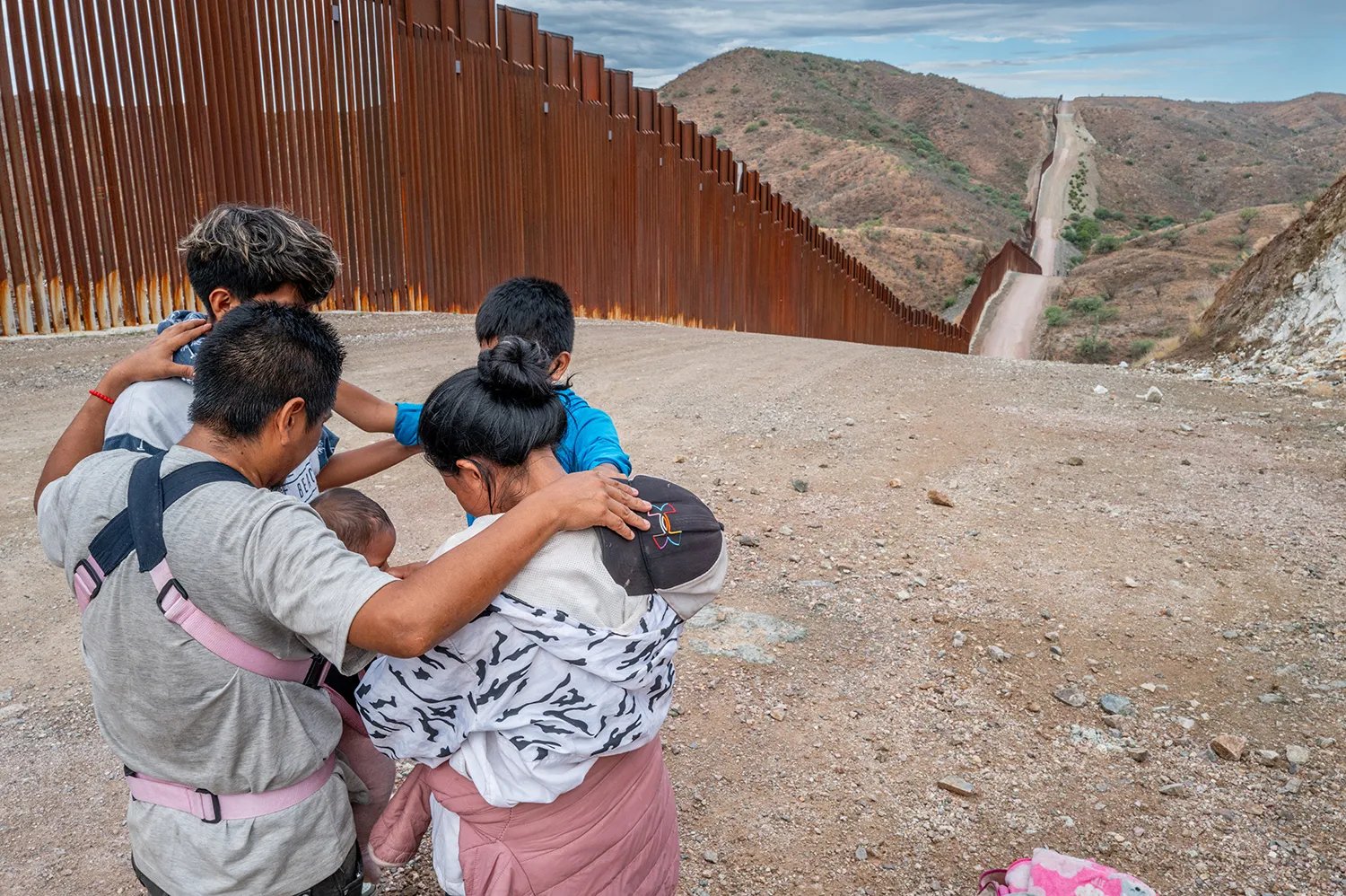 A five -year immigrant family, with a man, a woman, and a medium -class children, as well as a child in the transport company, puts their arms on the shoulders of each other while they stopped in a head circle. Behind them, long rusty metal barriers extend in the border fence along a rolling road on the side of the dust hill on the horizon.