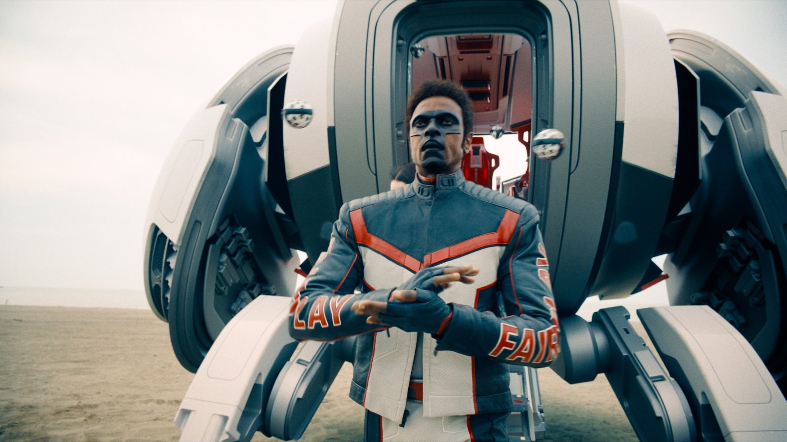 A man with a black mask in its shape like the letter T. The man is also a black, red and white motorcycle jacket with matching pants and standing in front of the Ferrean space ship.