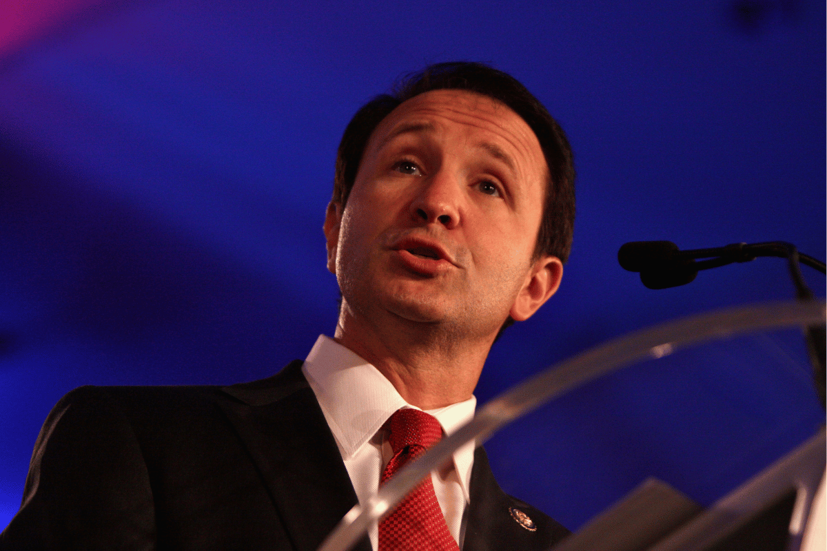 Jeff Landri's governor speaks on a platform platform, wearing a dark suit and red tie, with a microphone in front of him against a blue background.
