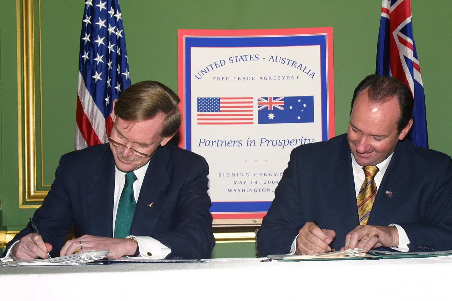 Two men sign documents before the flags of the United States and Australia as a poster reading "United States-Australia, Free Trade Agreement, Partners in Prosperity" appears behind them.