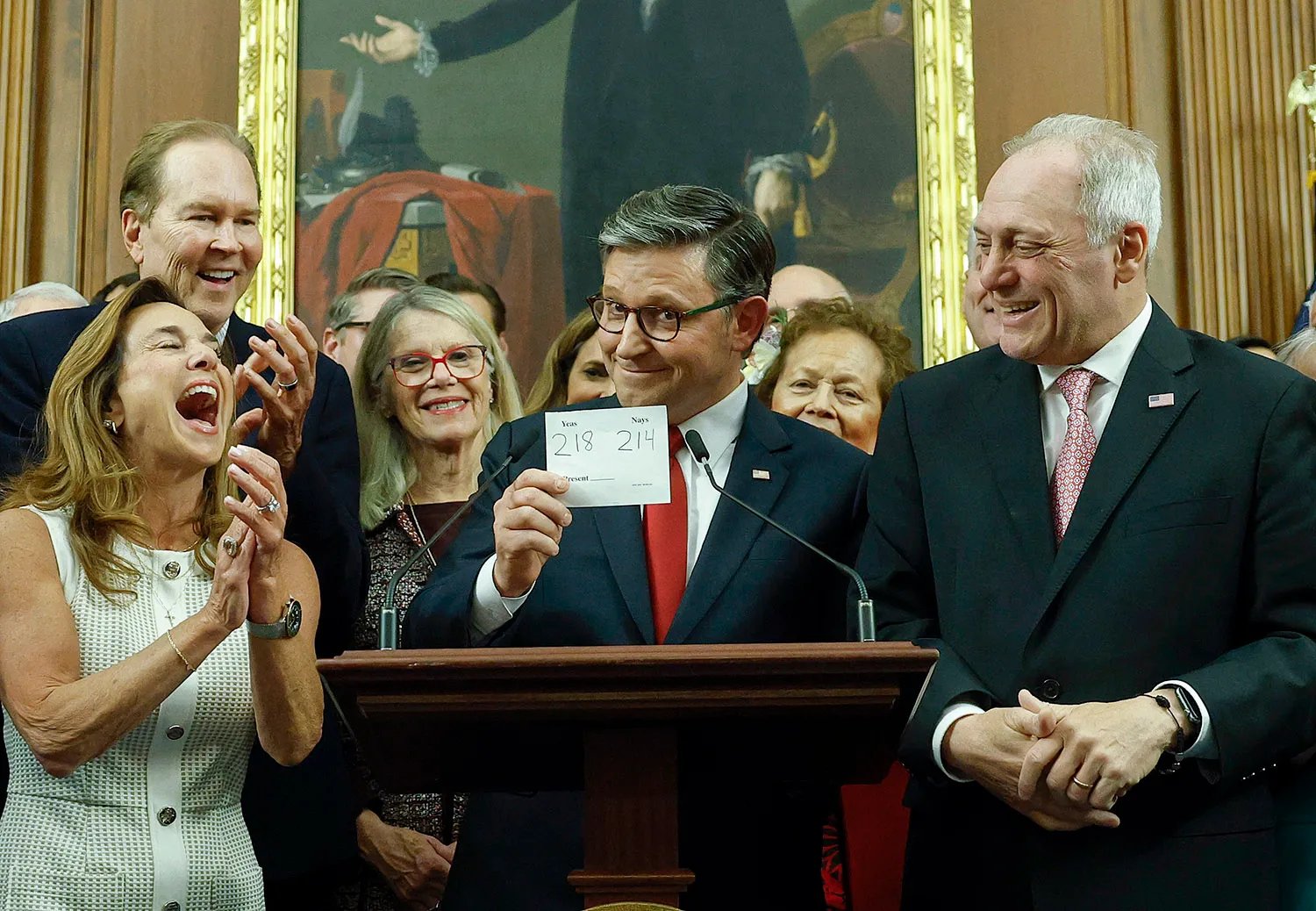 Mike Johnson stands behind a lectern and smirks at the camera while holding a 218/214 vote tally notecard. Around him is a cluster of about a dozen other lawmakers, who laugh and clap. A painting from the late 18th or early 19th century hangs behind them.