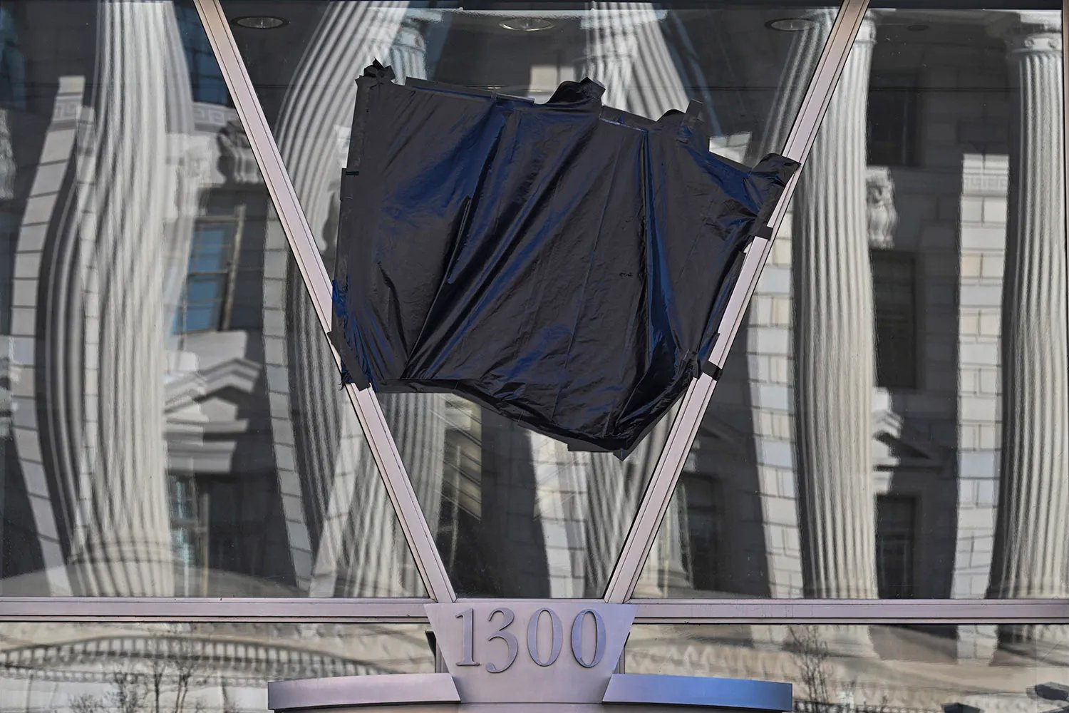 A black plastic rectangle over a sign on a window with twisted reflections of the columns fronting a large building.