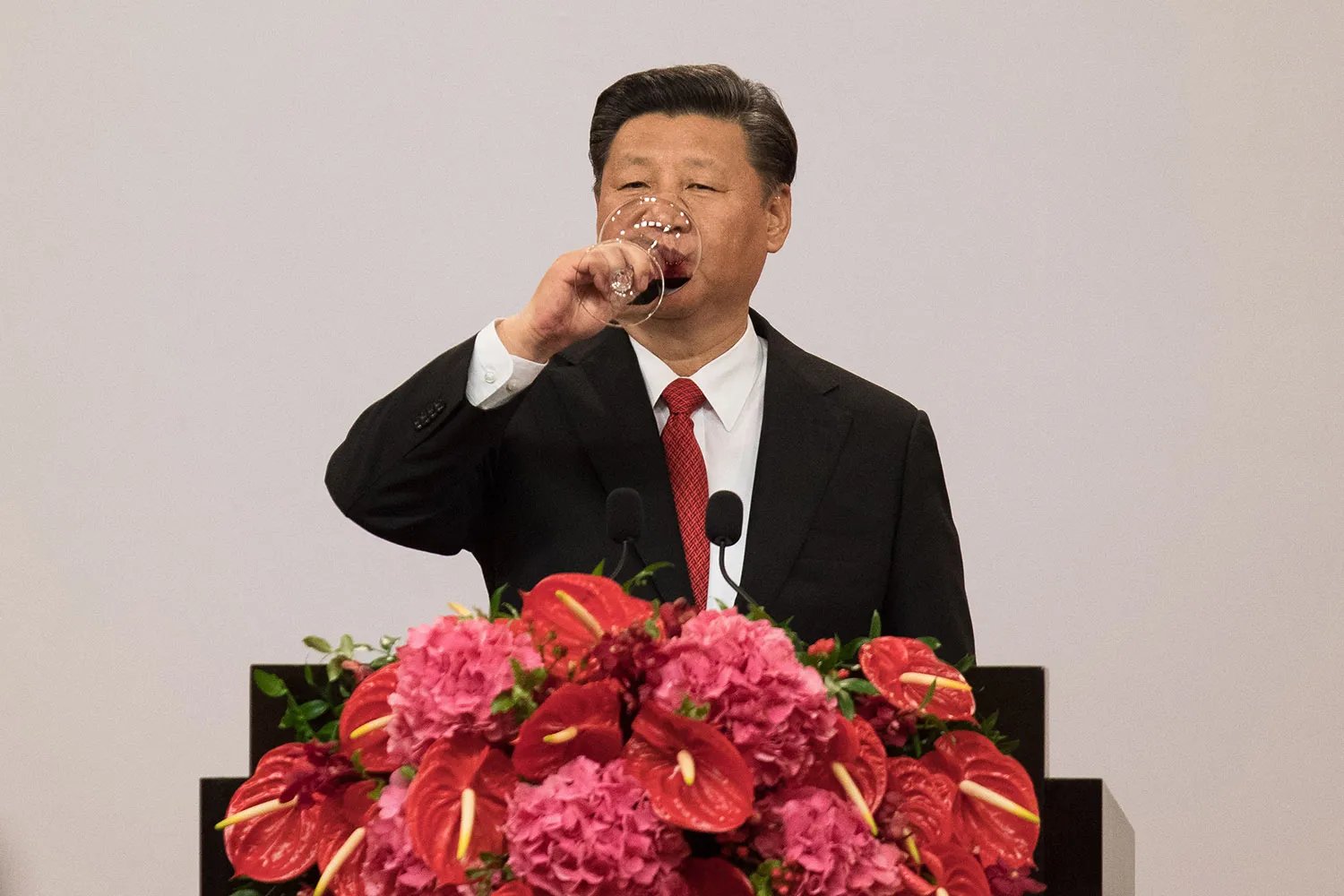 XI Jinping in a suit that drinks from a cup of wine behind flowers covered with flowers.