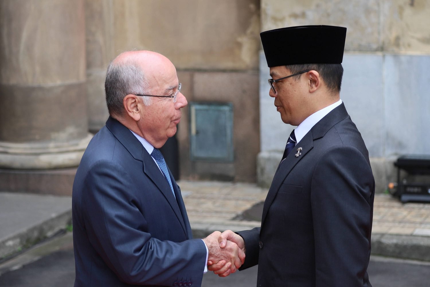 Two men wear suits, appear from the side, shaking hands.