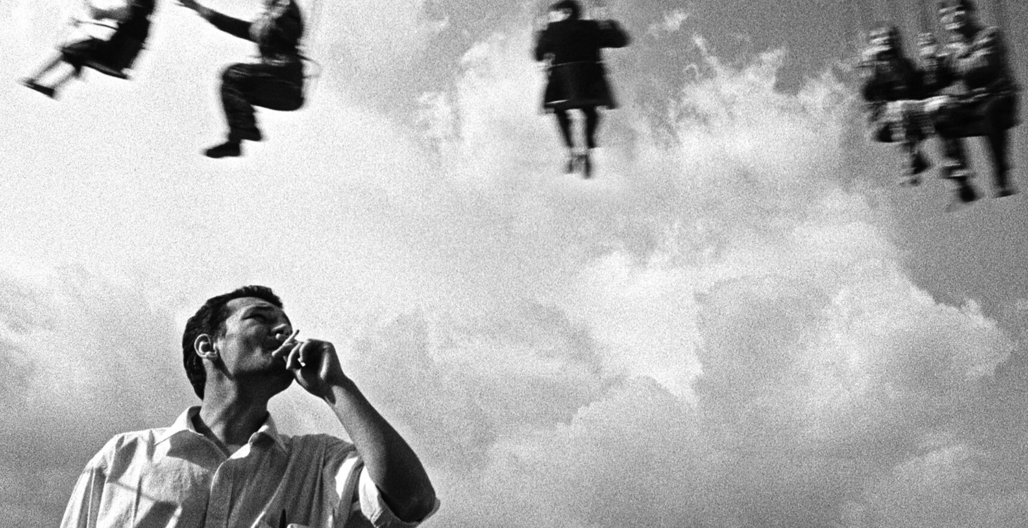 A man smokes as he looks up at a cloudy sky. The legs of people are seen swinging above him.