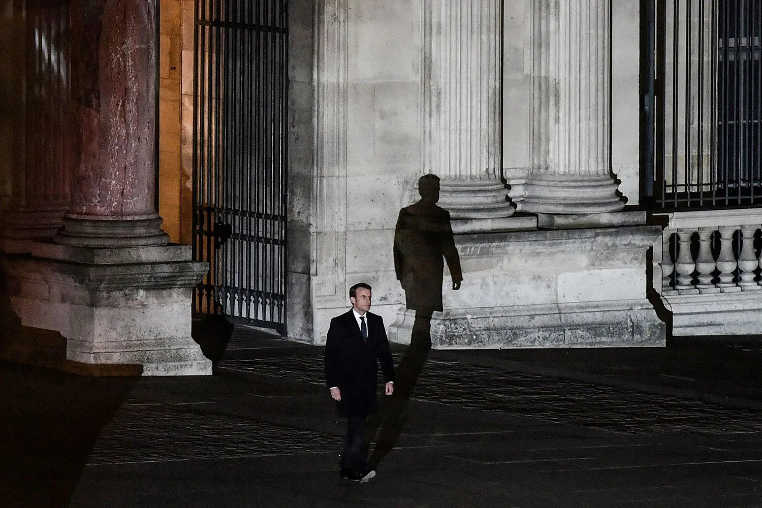 Macron casts a shadow on the exterior of a columned building as he walks forward into a courtyard.