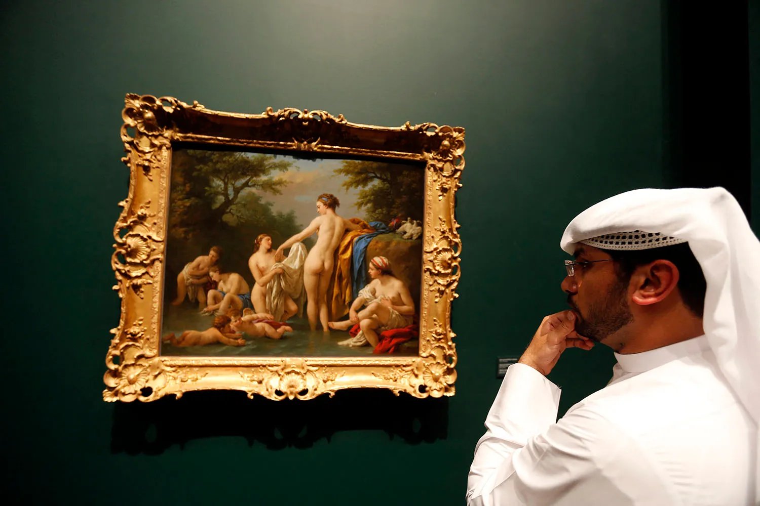A man wearing a head covering looks at a painting in a gilt frame showing nude women bathing.