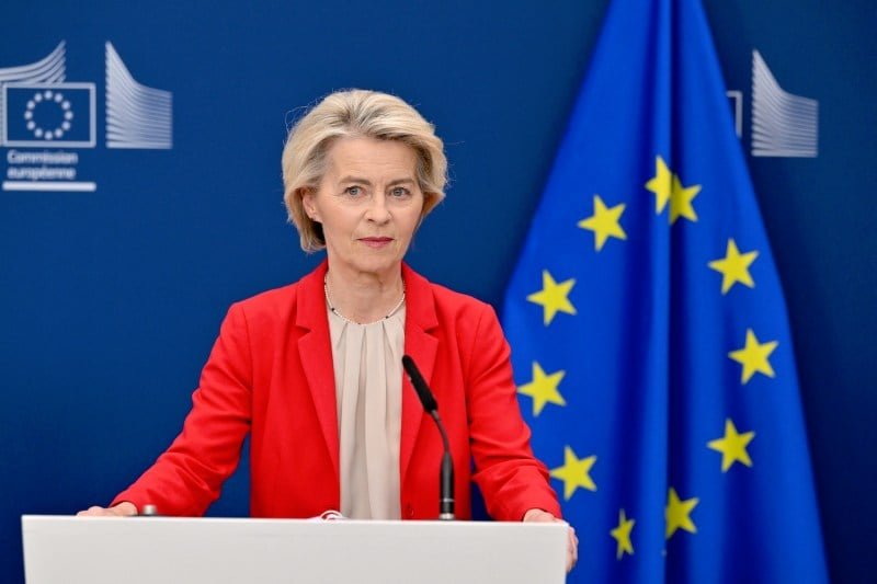 Ursula von der Lyen wears a beige blouse and a red laser while standing behind the platform. Behind it is the flag of the European Union.