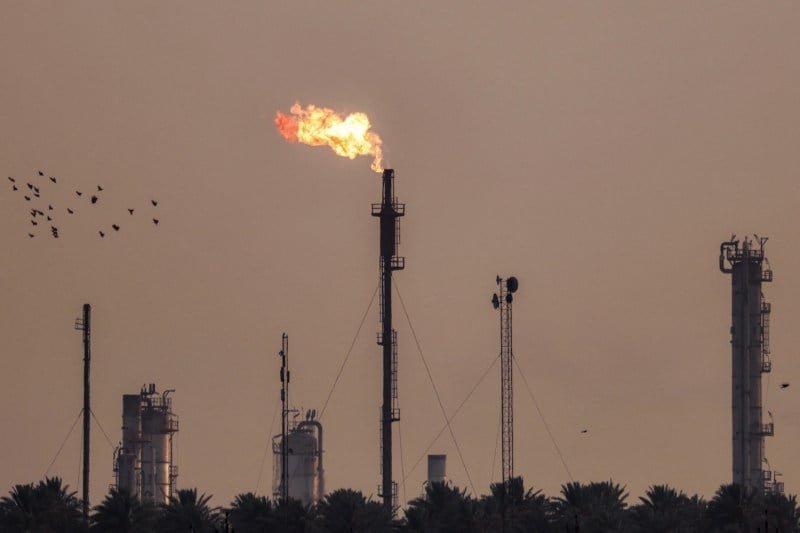 About twenty birds fly on the sky of blurry structure near the distillation and distress towers. The longest haven, in the middle of the image, has a pillar of flame that rises from above.
