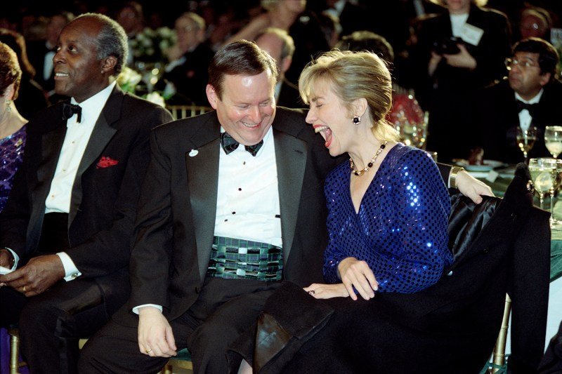 Al From in a tux with a green cumberbund laughs with Hillary Clinton, her mouth wide and eyes closed, and wearing a sequined top. Others in formal clothing are seated at tables around them.