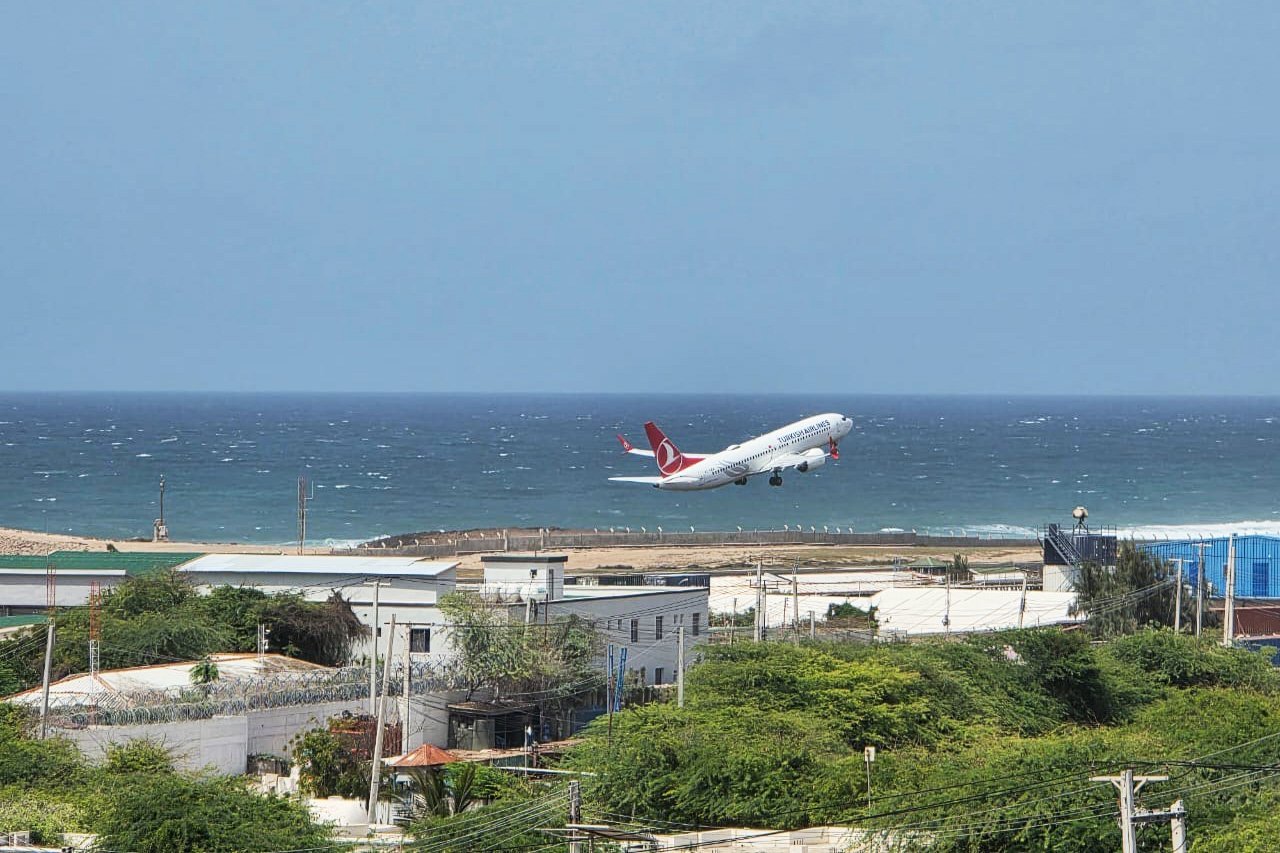 A plane is launched over the buildings and low trees along the coast of Mogadishu, with water on the horizon on the distance under the light blue sky.