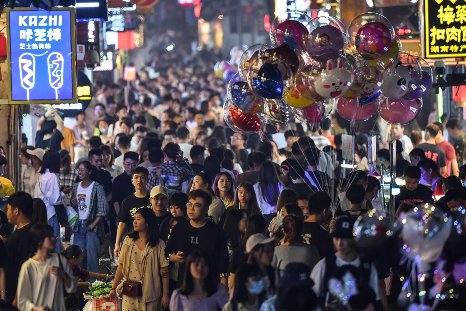 Pedestrians in a crowded street surrounded by small shops, with a person selling balloons in the foreground.