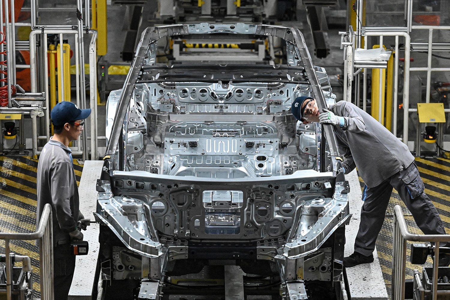 Two people on either side of a partially constructed electric vehicle on a production line. One person inspects the side of the car.